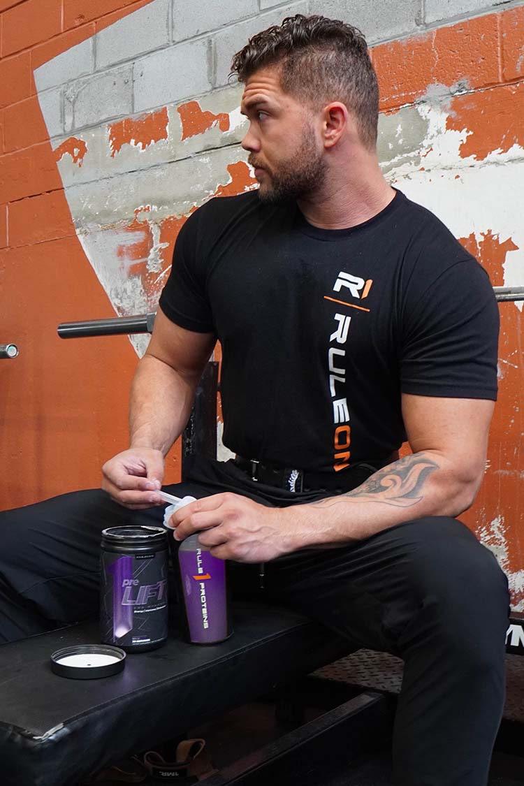 Man in a black 'Rule One' t-shirt sitting on a bench with supplement containers.
